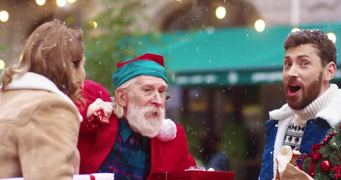Cheerful Santa Claus approaches beautiful young Caucasian couple and give them gifts. Surprised people become happy and cheerful at New Year's time.