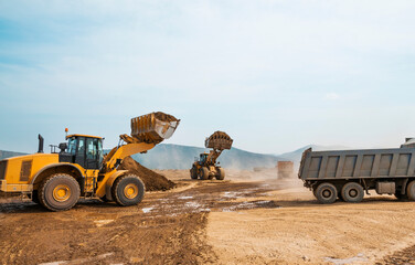 Earthworks on a summer day in a mountainous area. Wheel loaders, bulldozers and dump trucks are in operation. Dust from dump trucks that transport this soil