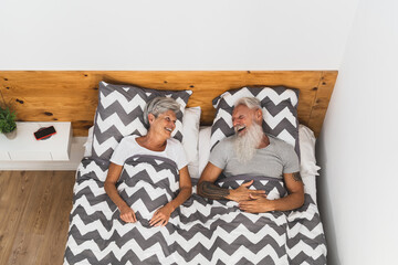 Happy senior couple smiling together in bed - Hipster mature people having funny bed time - Elderly lifestyle and love relationship concept