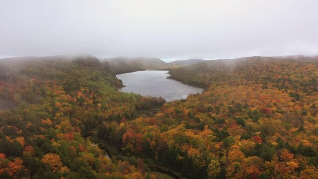 Beautiful panning out or back travel aerial of Lake of the Clouds and the pedestrian bridge over Carp River on an overcast morning with colorful autumn foliage blanketing the hills and valley below.