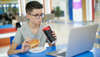 Teenage boy eating unhealthy fast food and drink soda while sitting in a food court