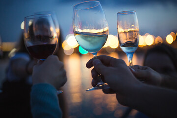 a party outside, celebrating and raising glasses with wine against blue night sky and yellow city lights, nice bokeh and lights reflections in the water - 395829520