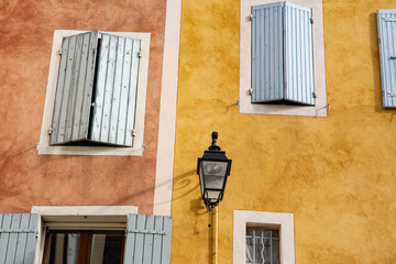 Facades of Provencal houses. One is yellow, the other is red, with white borders around the windows. Light blue shutters. A black steel lantern.
