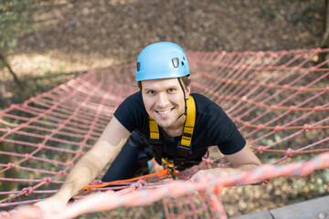 Well-equipped man climbing on a net outdoors, having active recreation in amusement park