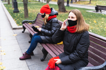 Fototapeta na wymiar Two young women, friends in face masks using, speaking mobile phone on safe social distance. Sitting on bench in autumn park.Meeting,talking during Covid-19 coronavirus pandemic.Warm jacket,red scarf