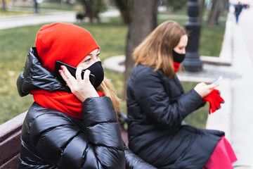 Two young women, friends in face masks using, speaking mobile phone on safe social distance. Sitting on bench in autumn park.Meeting,talking during Covid-19 coronavirus pandemic.Warm jacket,red scarf
