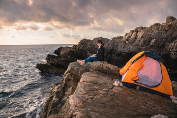 Fototapeta premium Female sitting on the rock above the sea and working on computer. Business woman working from the beach. Working from distance concept. Freelancer travel and work online.