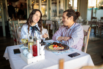 Fototapeta na wymiar Happy young mixed race couple enjoying dinner in a rustic restaurant, celebrating their anniversary or birthday. Looking at each other and talking