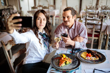 Happy young mixed ethnicity couple having dinner in a rustic restaurant, taking a selfie. Celebrating anniversary or birthday