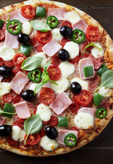 Pizza with ham, mozzarella cheese, cherry tomatoes, green and jalapeno pepper, black olives and fresh basil. Dark wooden background. Top view.	