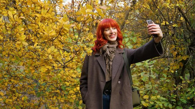 girl with red hair in a coat makes a selfie in the background of autumn foliage in the park