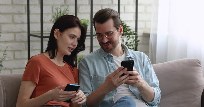 Addicted to technology happy young handsome man and beautiful woman sitting on sofa, sharing mobile devises usage experience, millennial couple spending free leisure time online, using smartphone apps