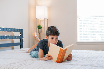 Adorable child boy laying on the bed and reading  a book, indoors. Child with book. Cozy home....