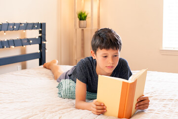 Adorable child boy laying on the bed and reading  a book, indoors. Child with book. Cozy home....