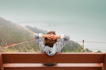 Back view of a girl in a gray hoodie. Woman sitting on a bench and admires the panorama of the city...