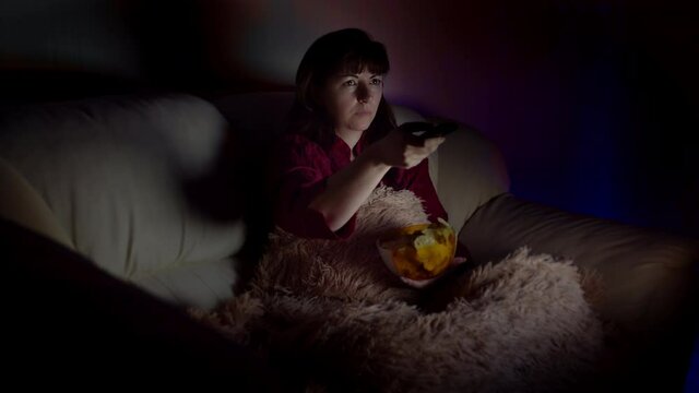 brunette woman sits in sofa with chips at night and watches tv with emotion