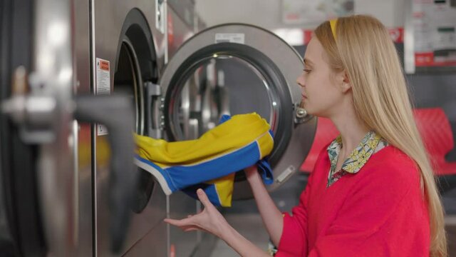 Young blond woman pulls out clothes the laundry into the washing machine from the basket in laundry room. Housewife domestic housework washer routine indoor. Slow motion