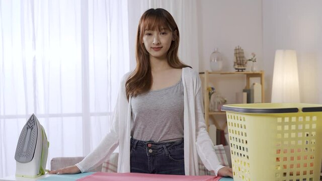 background bright home interior, beautiful asian female in casual wear standing behind ironing board with iron and laundry basket on it, looking at camera with a smile.