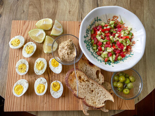 Above view of a Turkish breakfast platter. With halved boiled eggs, sourdough toast, a bowl of diced tomatoes and cucumbers,  halved lemons, a small glass bolw of hummus and a bowl of green olives