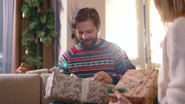 Excited caucasian bearded man receiving present from girlfriend opening gift box communicating spending winter holidats together. Christmas. New Year.