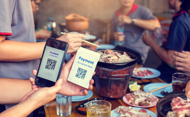 Hand holding smartphone to scan Qr code payment tag with blurry grilled pork and meat on the stove, Thai food buffet payment tag. QR code payment concept to many friends at a party in a  restaurant.