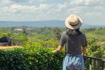 Fototapeta premium Asian woman wear weave hat and stand on wooden terrace. She feeling relax and happiness. she look at green trees and mountain hill.
