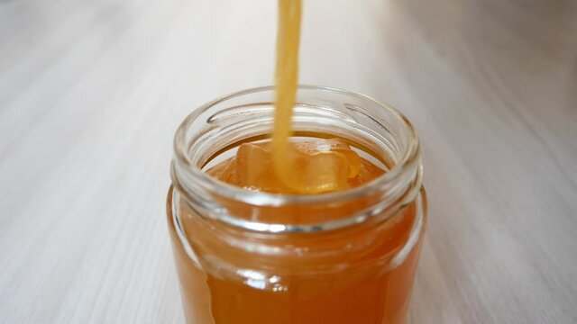 Fresh honey is poured into a glass jar on a wooden table close-up