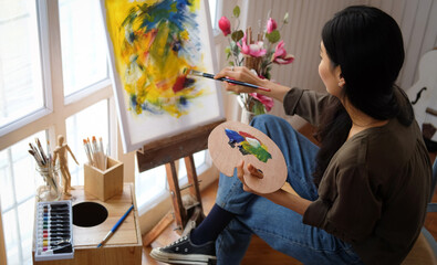 A young woman artist holding paintbrush and painting with oil paint on canvas at her workshop....