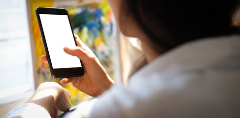Cropped shot of female artist using mobile phone with blank screen while sitting in front of the canvas painting in bright white studio.