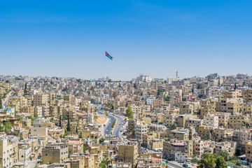 Cityscape of Amman with numerous buildings and Jordanian national flag, the capital and most...