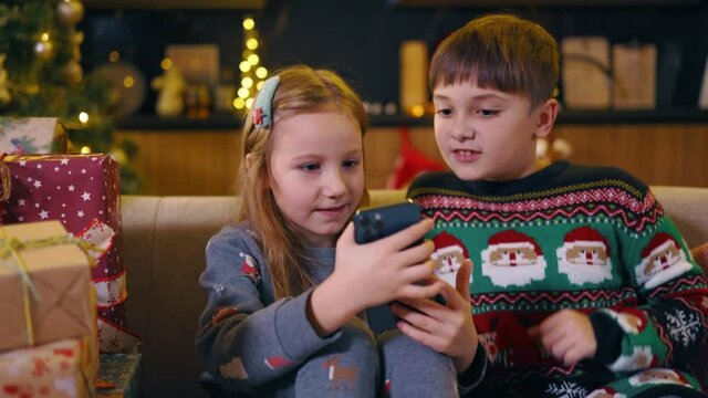 Couple of funny adorable children sitting on couch using smartphone application playing video games talking near Christmas decorated tree. Winter holidays.