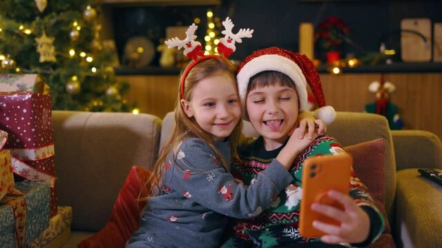 Couple of cute adorable children wearing xmas hats having fun using smartphone taking selfie picture near Christmas tree. Winter season. Holidays and kids.