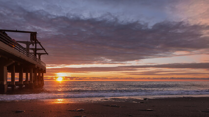 Evening. The sun shines low over the sea. Blue clouds are highlighted in pink. There is a sunny path on the water. Waves foaming on a pebble beach. On the left is an empty pier. Fall. Russia