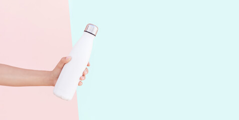 Close-up of female hand holding reusable steel thermo water bottle of white. Panoramic background of pastel pink and cyan of colors with copy space.