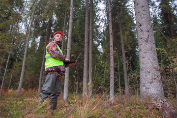 A forest engineer in a helmet and a vest works in the forest with a computer. The concept of computerized forest inventory.