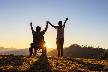Fototapeta na wymiar Disabled handicapped young man in wheelchair raised hands with his care helper in sunset.Silhouette