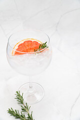 A single large bowl style glass of Gin and Tonic served with ice, pink grapefruit and rosemary. Glass filled with ice and liquid against white marble texture background. Copy space.