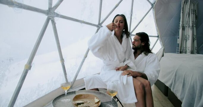 Woman sitting on a man's knee eating, in a white bathrobe and kissing. Attractive man and woman chilling in an igloo hotel room 4K