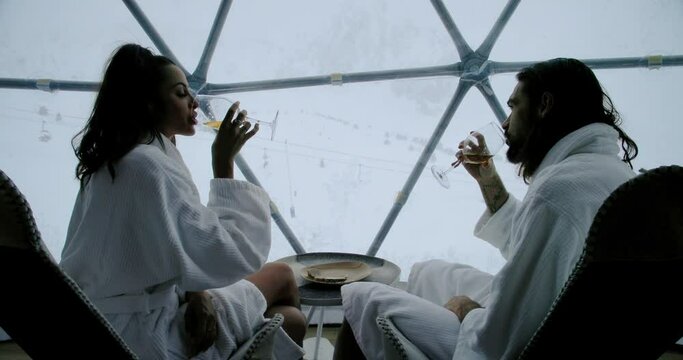 Woman sitting on a man's knee drinking white wine, in a white bathrobe and look at a ski station. Attractive man and woman chilling in an igloo hotel room - behind view 4K