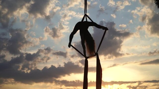 Silhouette of Female circus artist showing her flexibility and splits with red aerial silk on the dramatic sky background in slow motion. Concept of healthy lifestyle 