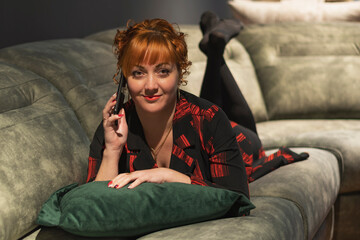 Obraz premium A young red-haired girl lies on a sofa in a dark room, holds a mobile phone to her head and listens attentively to the conversation on the phone
