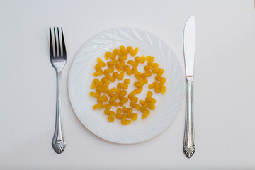 On a white plate, scattered raw dry yellow spun pasta, spiral, next to a fork and knife. White...