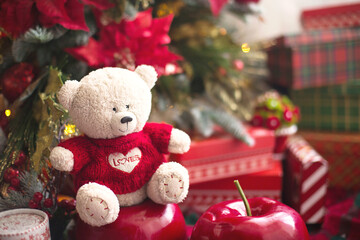 White Teddy bear in red knitted sweater with heart on the chest and the words Love near Christmas tree among the gift boxes. Gift to your beloved, Declaration of love, poinsettia decor. Copy space