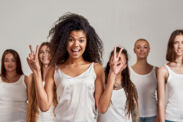 Fototapeta premium Portrait of young african american woman in white shirt showing peace sign at camera. Group of diverse women standing isolated over grey background