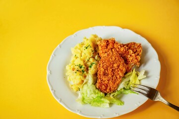 Chicken piece breaded in cornflake, mashed potato and salad in plate, fork on yellow background.