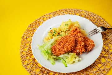 Chicken piece breaded in cornflake, mashed potato and salad in plate, fork on yellow background.