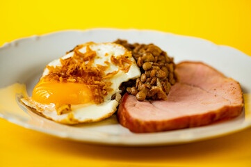 Fried egg with onion, boiled lentil and sliced smoked meat on white plate on yellow background.