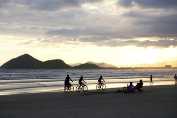 People riding a bicycle on the beach sand at dusk. Family exercising and other resting in chairs.