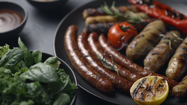 A dish of grilled sausages with smoke, tomatoes, lemon, vegetables. Fresh salad, dark background. The camera moves to the dish