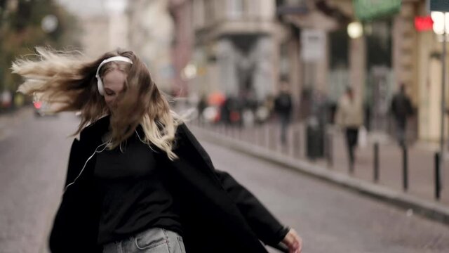 Happy young woman in headphones dancing outdoors in city street having fun alone. Joyful attractive blonde carefree woman listening to music with smartphone